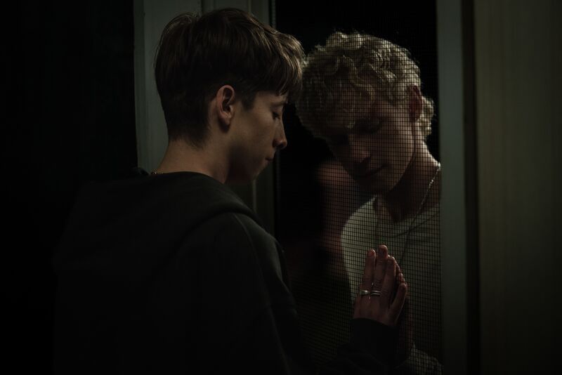 Two young men lean their heads together and press hands through a mesh door