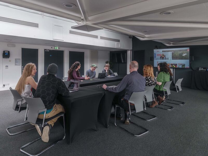 A group of people sit around a u-shaped table in an office meeting room having a conversation. There is a screen visible in the background