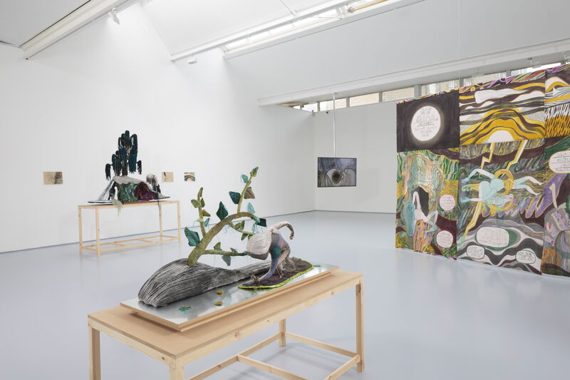 A hung textile work with watercolour painting and graphic text hangs to the right. A monitor hangs from the ceiling to the back right. In the foreground right is a plinth with a textile piece and another can be seen behind it on the far right. The colours are muted and soft, the gallery is naturally lit and bright. 