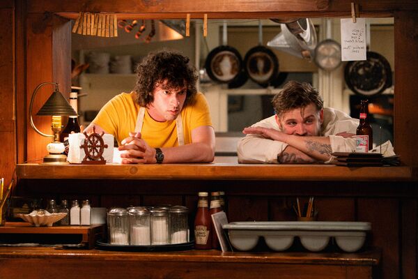 Two young chefs look into a restaurant from the kitchen window