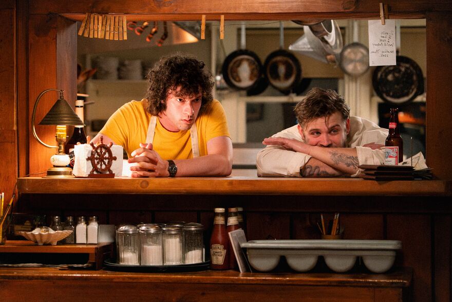 Two young chefs look into a restaurant from the kitchen window