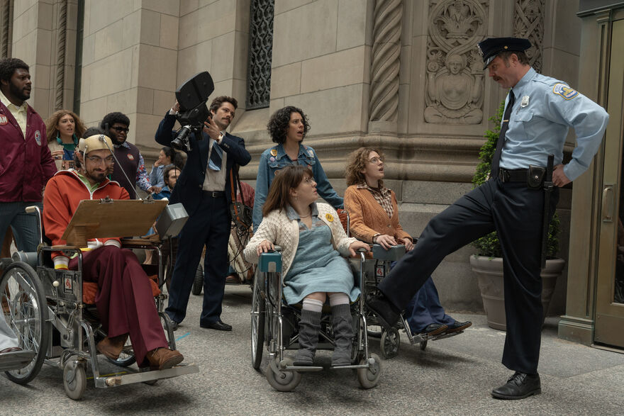 A policeman puts his foot on a woman's wheelchair while people look on appalled