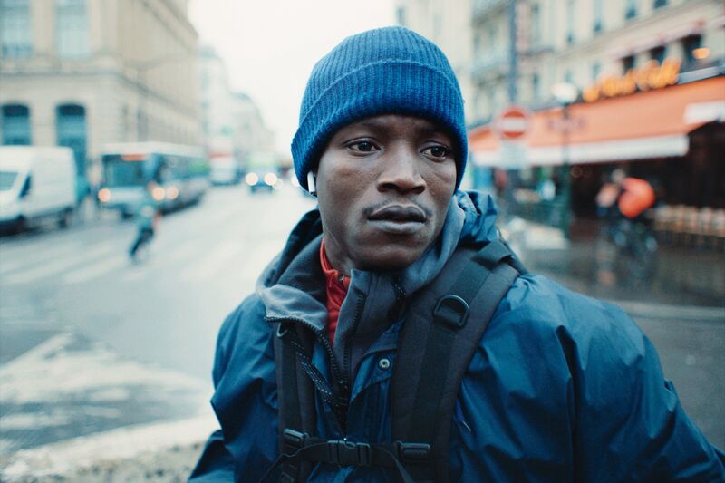 A young man wearing a blue hat and airpods stands in a rainy street in Paris.