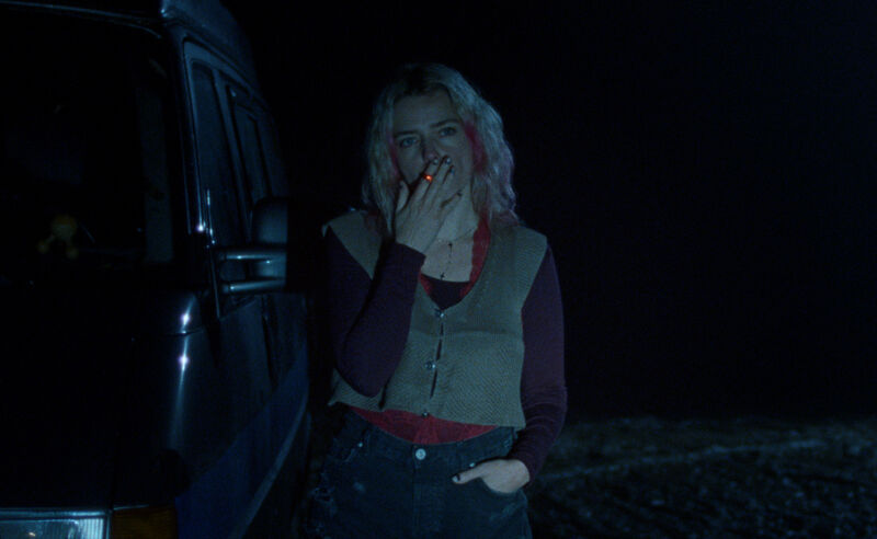 Blonde woman standing beside a vehicle smoking