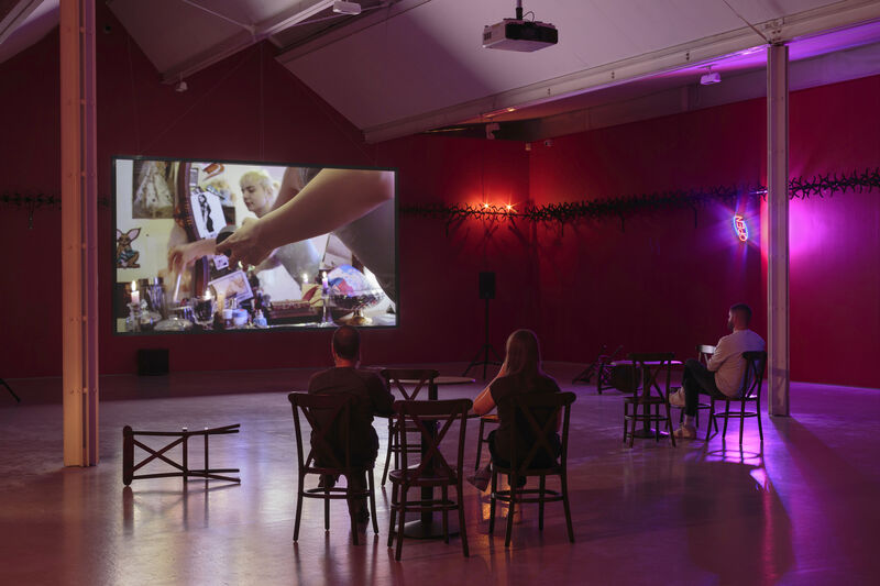A wide shot photograph of gallery two, showing the gallery with red walls and cabaret-style seating with small tables in the floorspace. A few people sit and face a screen with a projected film. There is a bright purple light to the back right of the image.  