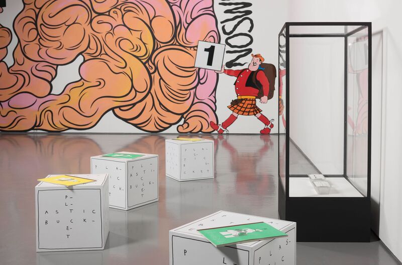 In front of a giant mural painted on the wall which shows Desperate Dan, there is a transparent box with two small foil takeaway boxes inside them. Next to the box are four smaller white boxes with the word 'Plastic Bucket' written on them. On top of these boxes are green and yellow books.