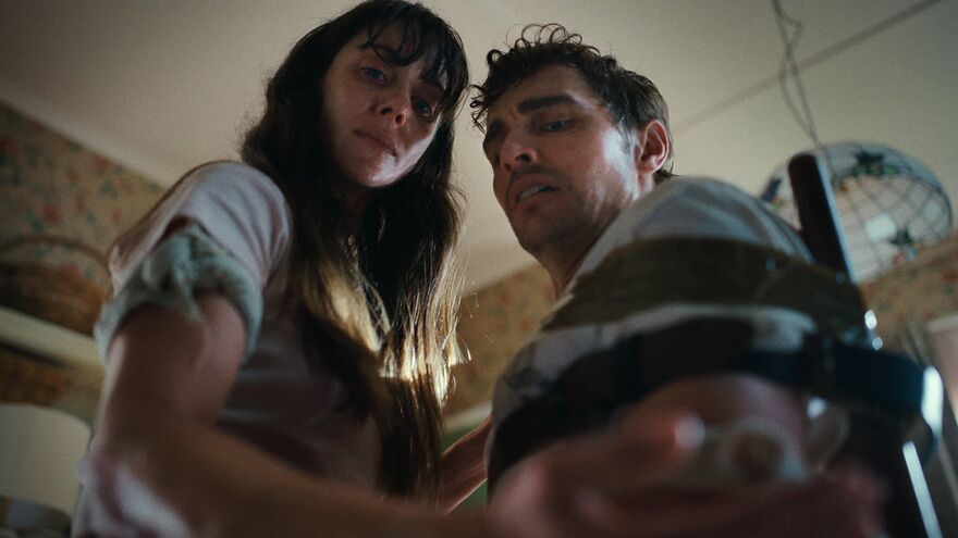 A man and woman stand next to each other, covered in blood and sweat. The man is tied to a chair.