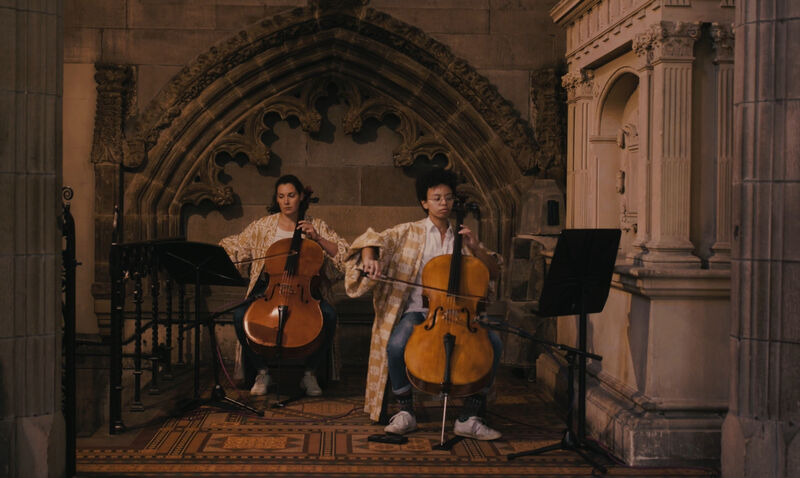Two people playing the cello in a church like space 