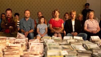 A row of serious-looking people in casual clothes sit in front of a giant pile of bank notes.