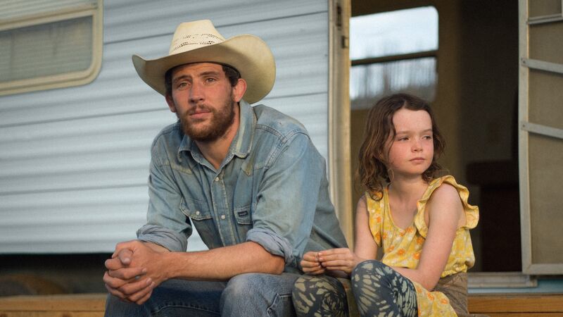A man wearing a cowboy hat and his daughter sit on the step outside their trailer home