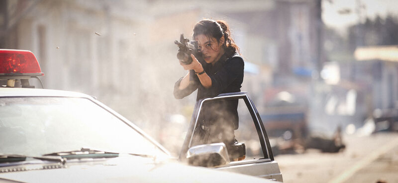 A woman aims a rifle from behind a police car