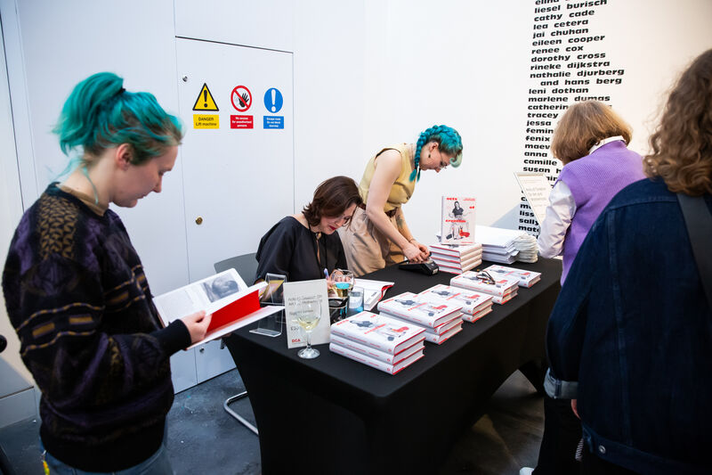 A desk with books on it with two women behind the desk one writing in the book the other standing 