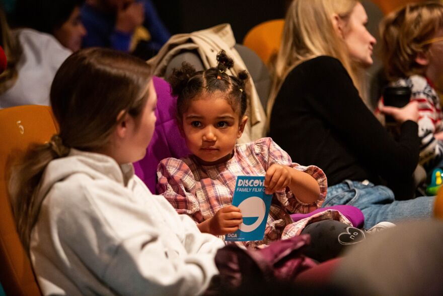 Two girls chatting at DCA cinema. One of them holds a Discovery Family Film Club flyer.