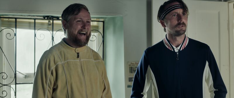 Two men dressed in athletic clothes stand next to each other. One of them laughs while the other looks pertubed.