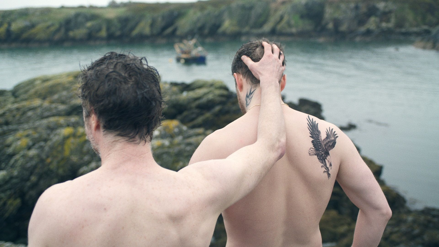 Two young men stand on cliffs above the sea, ready to dive and swim