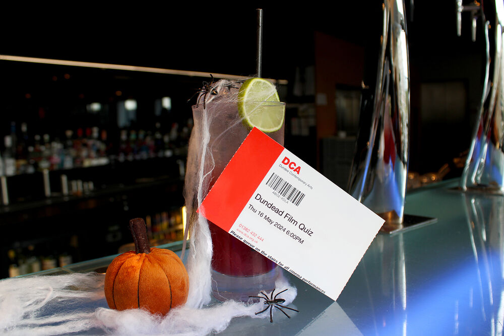 Dundead Film Quiz ticket on teh bar beside a drink, fake spiders and cobwebs and a small plush pumpkin