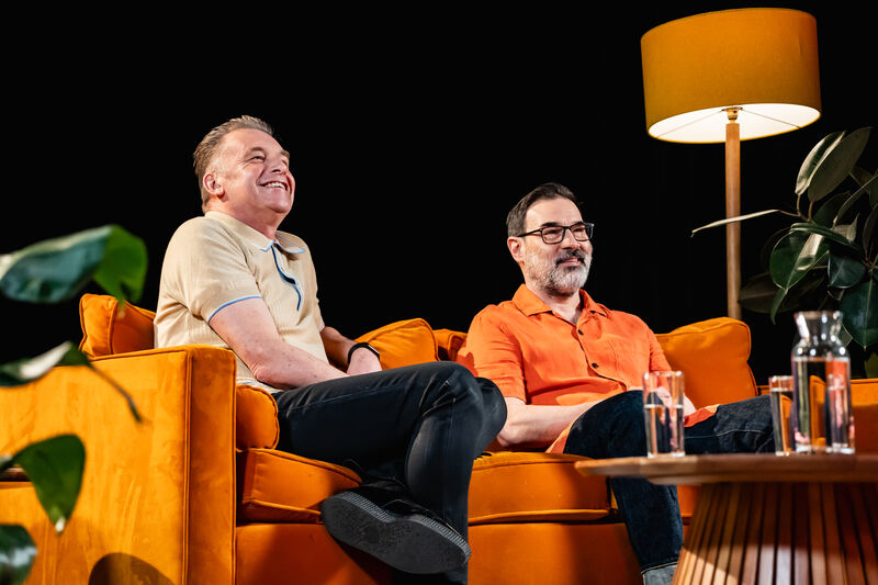 two men sit on an orange sofa looking at the camera