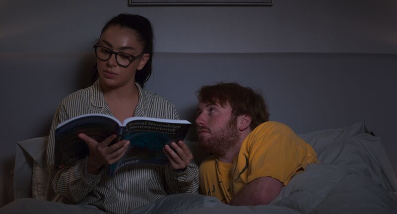 A young woman reads in bed while a young man stares up at her