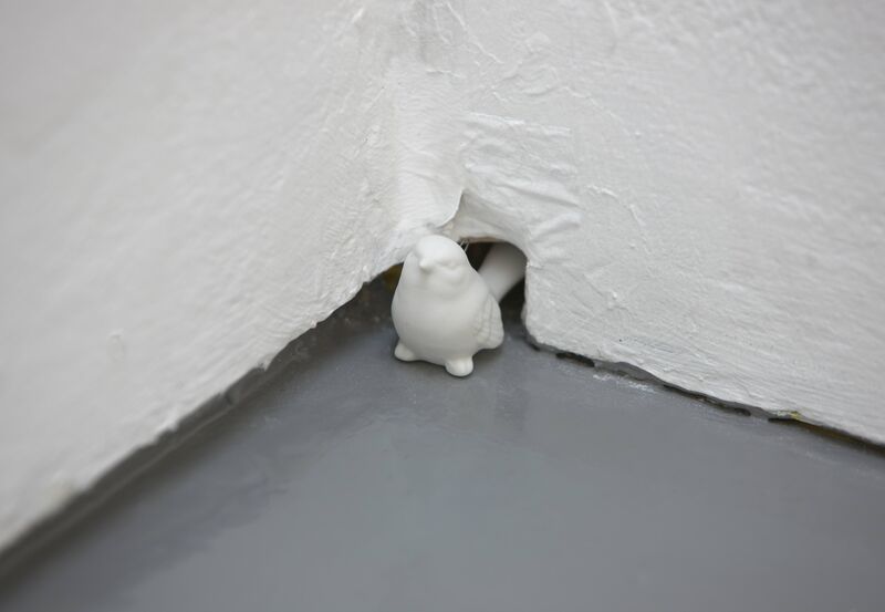 A small porcelain figure of a bird stands next to a small hole in the corner of a wall in Johanna Basford's exhibition at DCA.