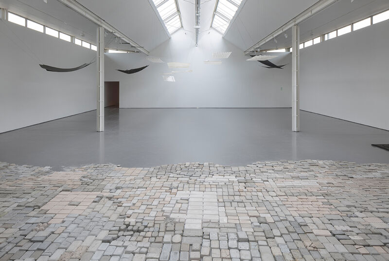 A wide installation photograph showing gallery 2, with many cast brick-like objects laid on the floor in a grid in the foreground, and a series of works suspended from the ceiling consisting of black and white metal sheets. 