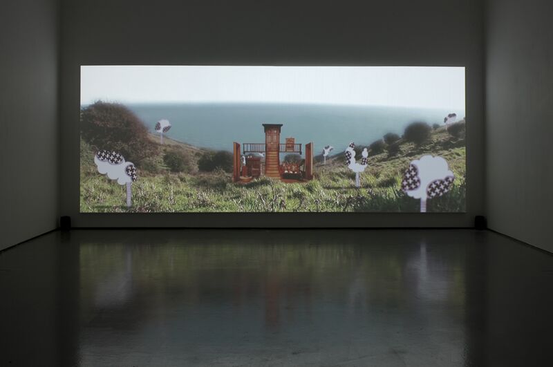 From Hiraki Sawa's exhibition. A screen shows A model of a tiny living room sits on a grassy cliff edge, with the sea in the background.
