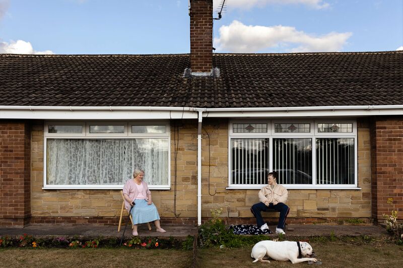 An older woman and a younger woman wearing a tracksuit sit outside of their bungalows together