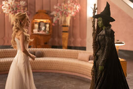 A blonde woman in a white dress and green skinned woman wearing a witch's hat an holding a broom stick stare at each other.
