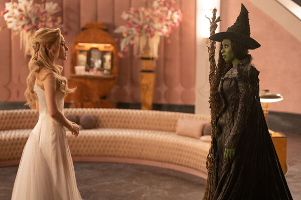 A blonde woman in a white dress and green skinned woman wearing a witch's hat an holding a broom stick stare at each other.