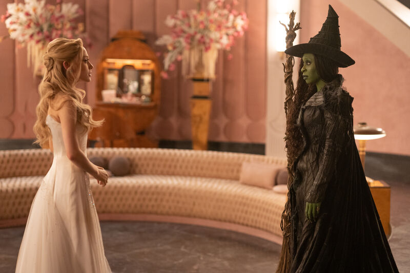 A blonde woman in a white dress and green skinned woman wearing a witch's hat an holding a broom stick stare at each other.
