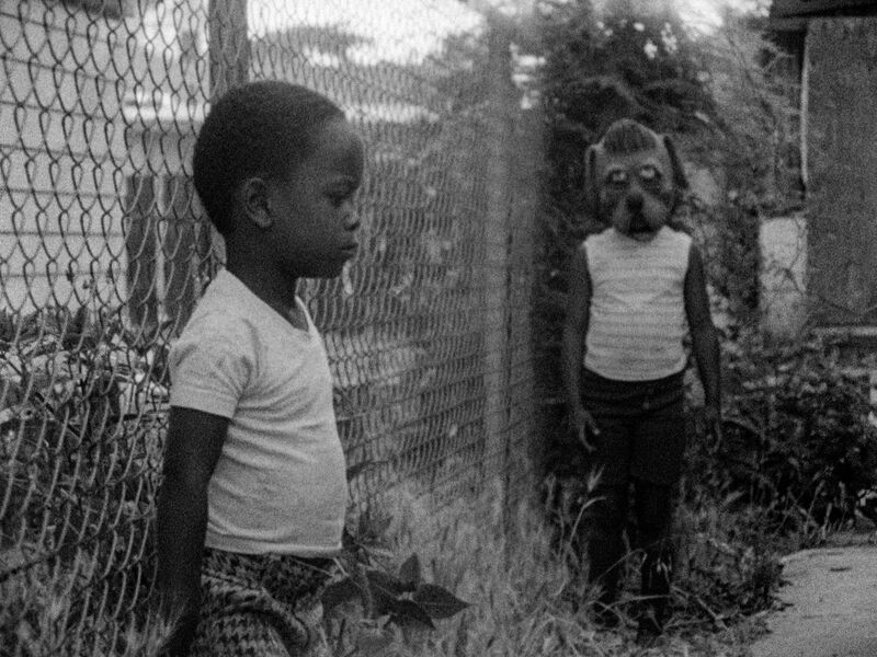 a black and white image of a young black boy standing against a fence with another boy wearing a dog mask in the background