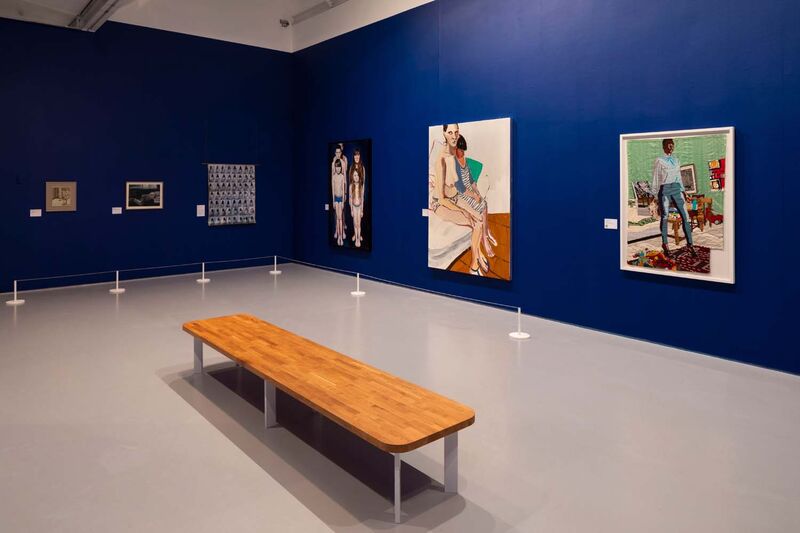 The corner of  a blue gallery with large paintings hung on it. A bench is in the centre 
