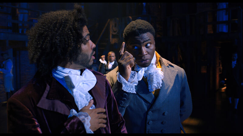 A man in 18th century clothing looks at another man and points.
