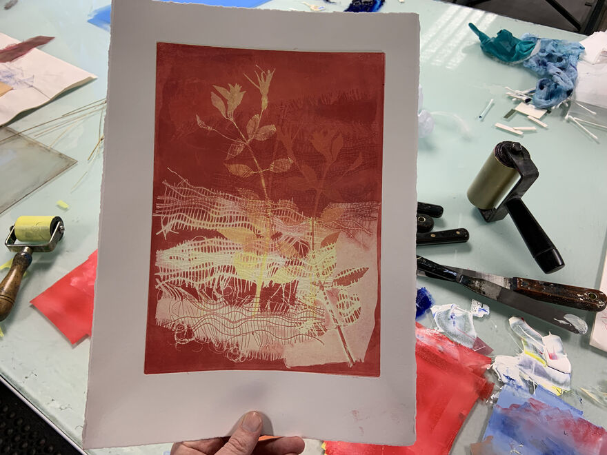 Person holding up a mono print of plants in front of a desk covered in mono printing ink and equipment