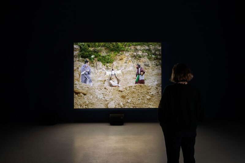 This image shows a dark gallery with a video projection. In the film we can see three figures in traditional Japanese clothes, posed in a quarry-like setting with loose pale rocks and weedy plants. 
