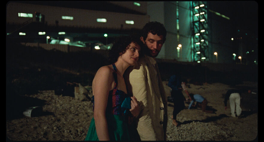 Two people walk outside at night. A large building is lit up in the background. The two people are linking arms.