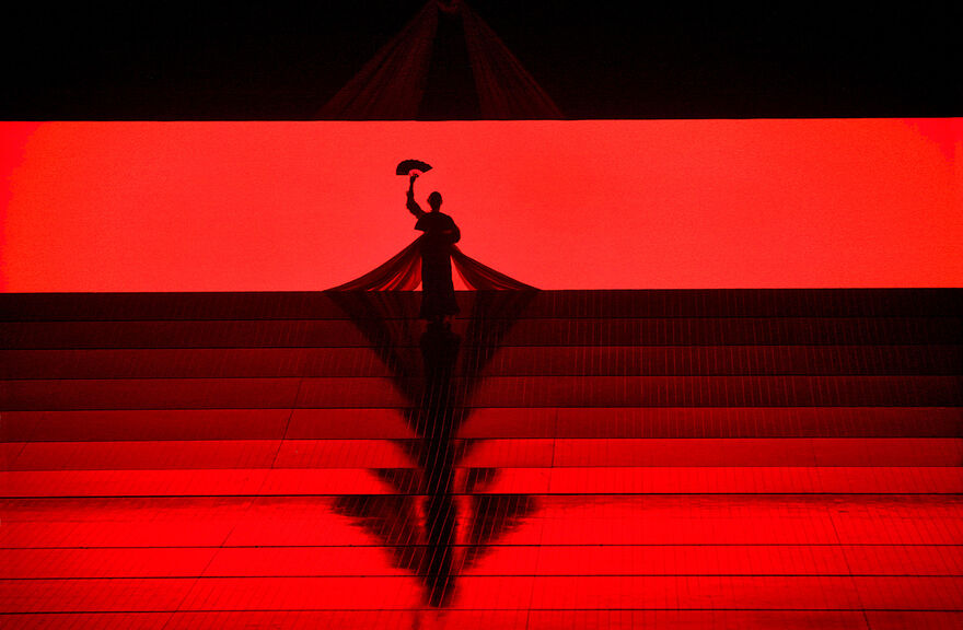 A scene from Madama Butterfly shows a silhouette of a figure holding a fan against a red background.