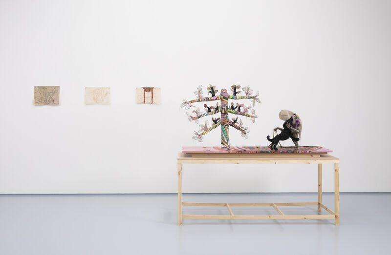 Three small works on paper can be seen on a white wall to the left of the image. To the right is a wooden plinth with a textile sculpture. In the sculpture is a figure with their head bend to the left, and on the right is a tree-like form with six lumpy branches. The colours are mainly muted pinks, yellows and greens. 
