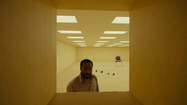a man looks through a window in a strange basement room, with empty shoes and a chair sunk into the floor.