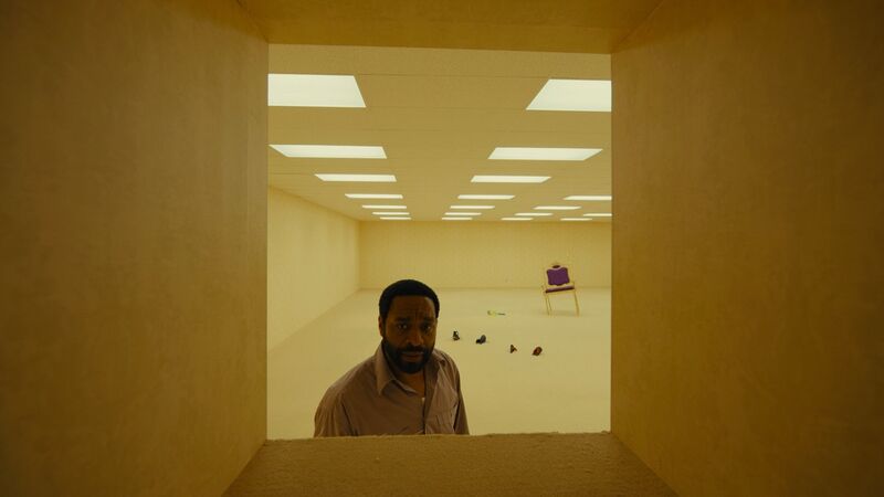 a man looks through a window in a strange basement room, with empty shoes and a chair sunk into the floor.