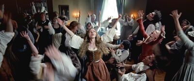 A woman in medieval clothes dances wildly in a room of other people who are also rhapsodising and dancing
