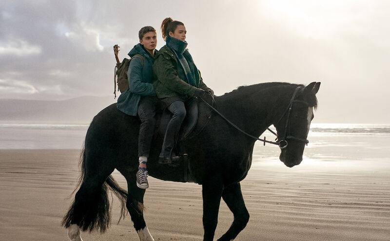 Two people ride a large black horse along a sandy beach, with the sea and cloudy sky visible in the background.