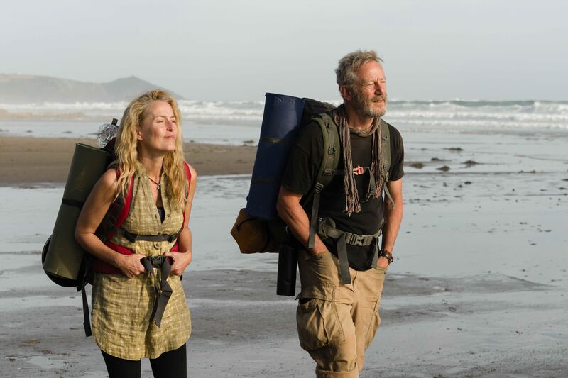 A middle-aged man and woman wearing back packs walk across a beach.