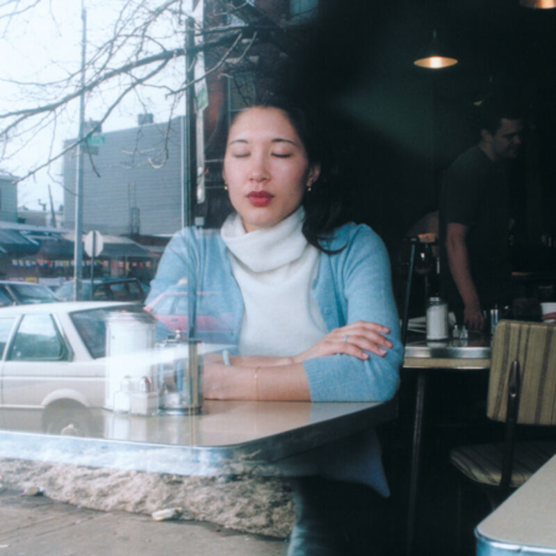 From Banner/Lislegaard's exhibition. A photograph of a woman with her eyes closed, sitting in a cafe. Out the window, is a city scene with a car and bare trees.