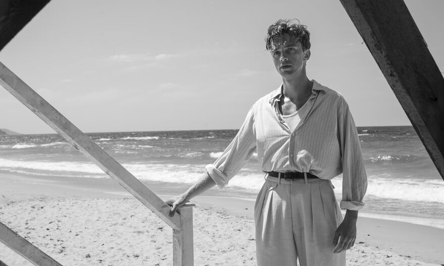 A black and white image of a man standing at the bottom of some wooden steps on a beach with the sea behind him