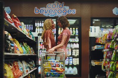 Two women soaked in blood stand in a supermarket looking at each other and holding a trolley