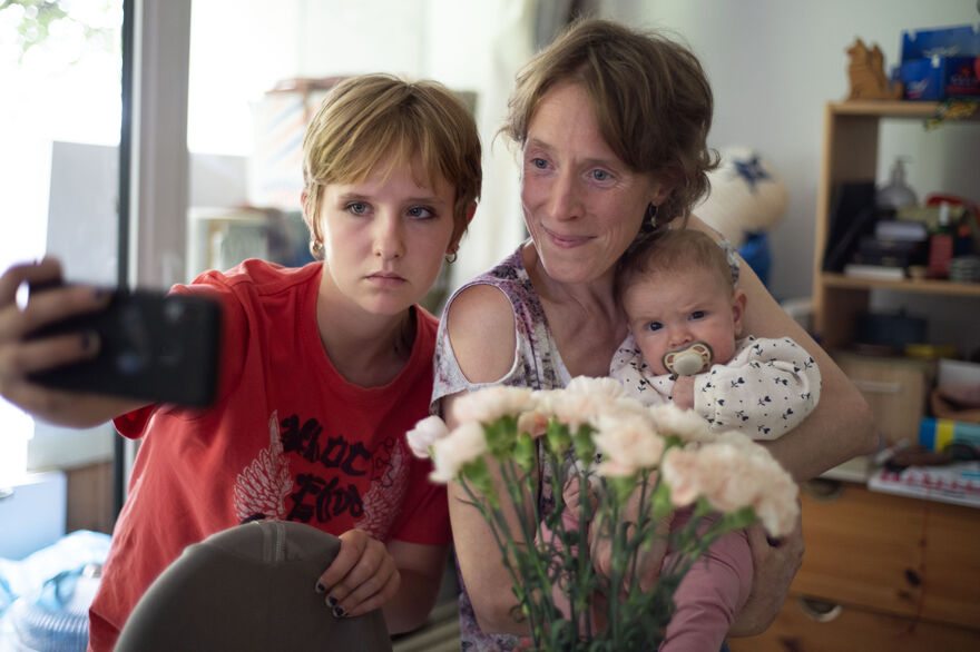 A young and an older lady take a selfie with a phone while holding a small baby with a dummy in its mouth