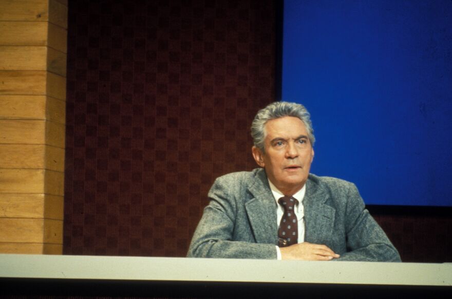 A person in a grey suit sits behind a desk on a television studio set, with wood-panelled walls and a large blue screen in the background.