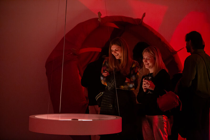 Two people smile and hold glasses of wine as they look at Michelle William's Gamaker's work in a hanging round vitrine. 
