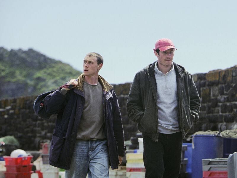 Two men walk through a fishing village.