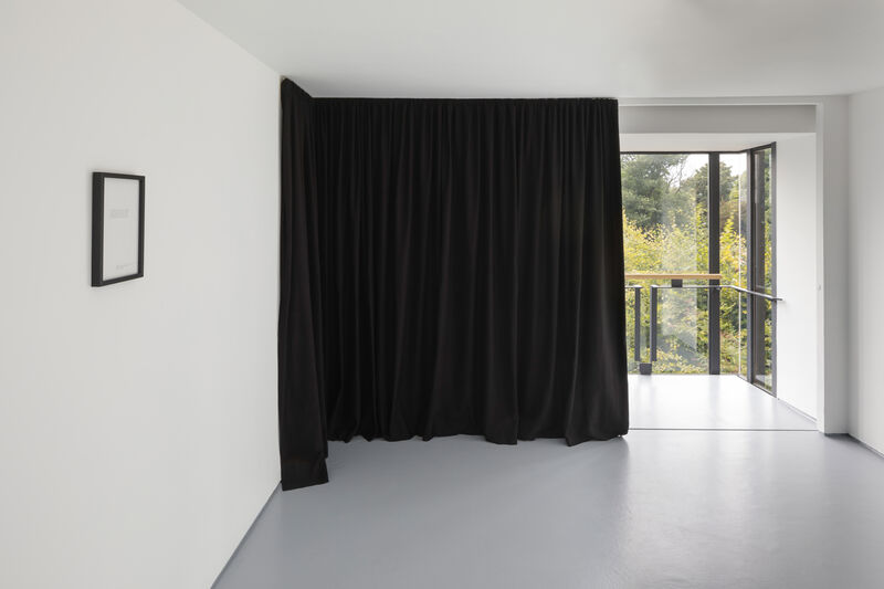 The is a photograph of the auxiliary gallery at the back of gallery two, showing a large black corner in the left hand corner, partially covering the window and the left hand wall. To the far left of the photograph we can see a small framed artwork on paper. 